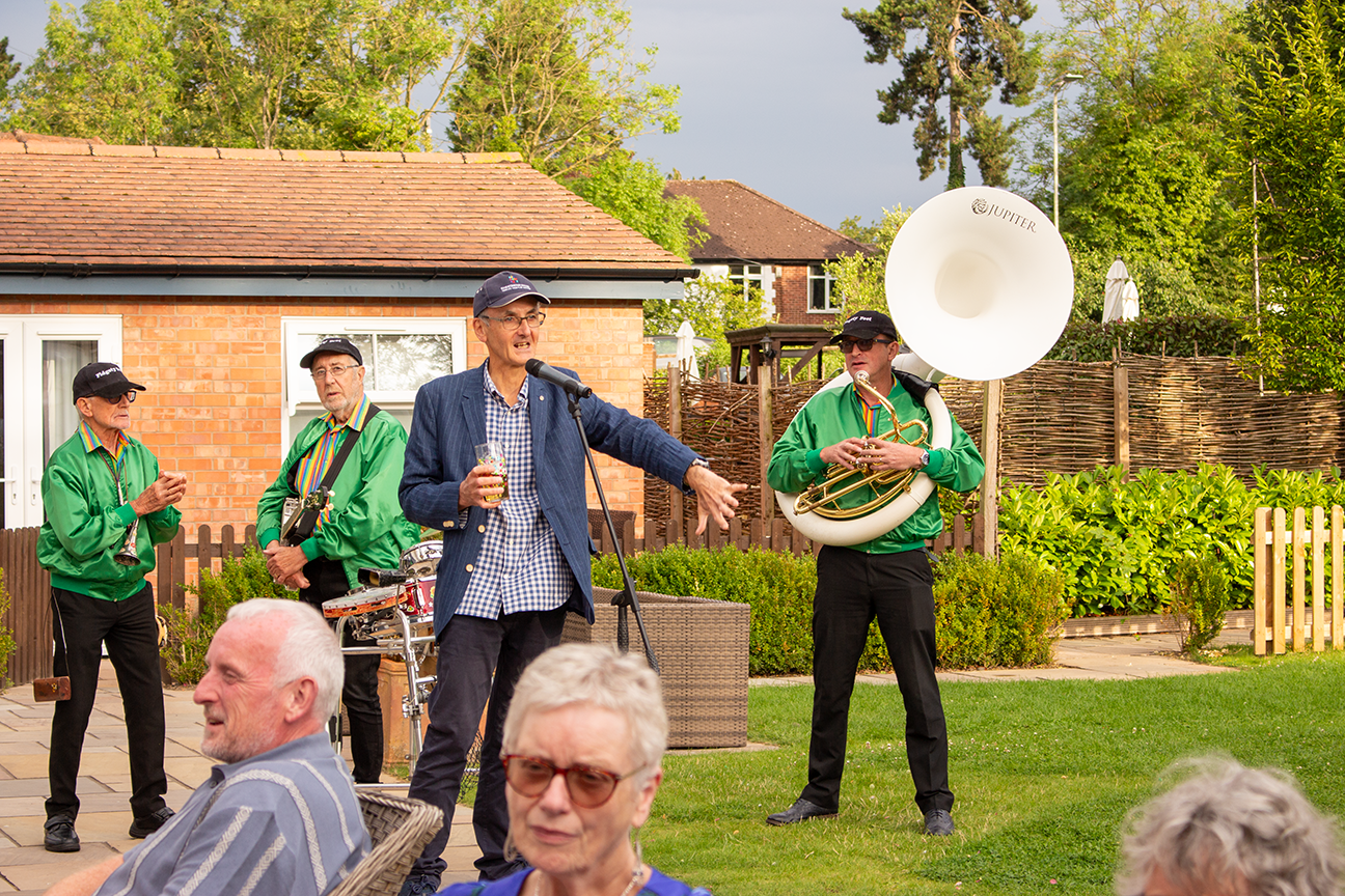 Andy Jervis stands speaking in the venue garden with the band behind him on a summer evening