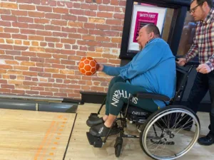 Headway Leicester. Image of man in wheelchair holding an orange ball at the start of a bowling lane.
