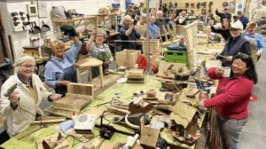 Men and Women in Sheds Charnwood. Image of a group of people around a workbench with woodwork and tools in front of them. Taken from bbc.co.uk.