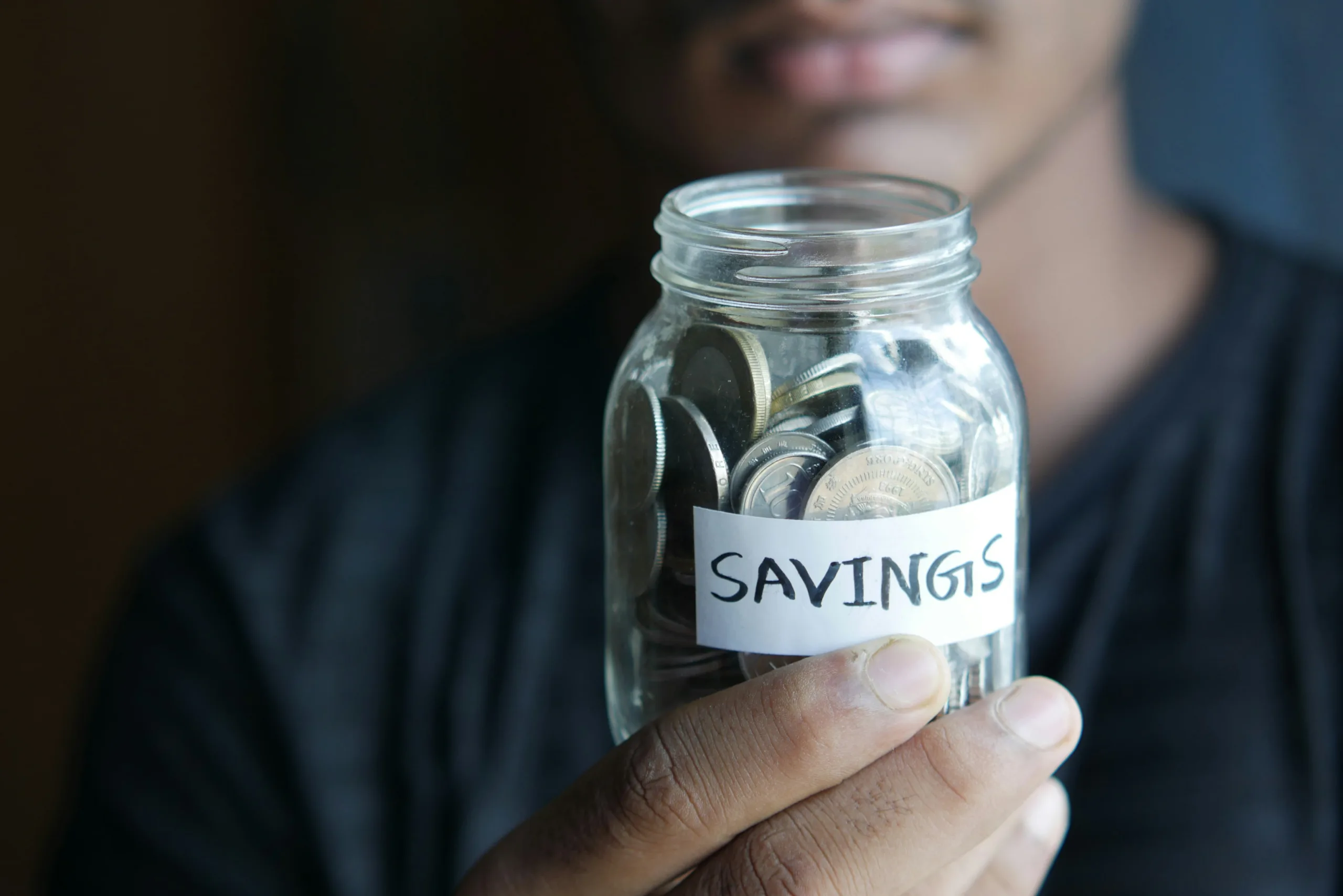 Decorative image to accompany blog about savings. Man in background blurred, holding a glass jar containing coins and a lable on the front saying savings
