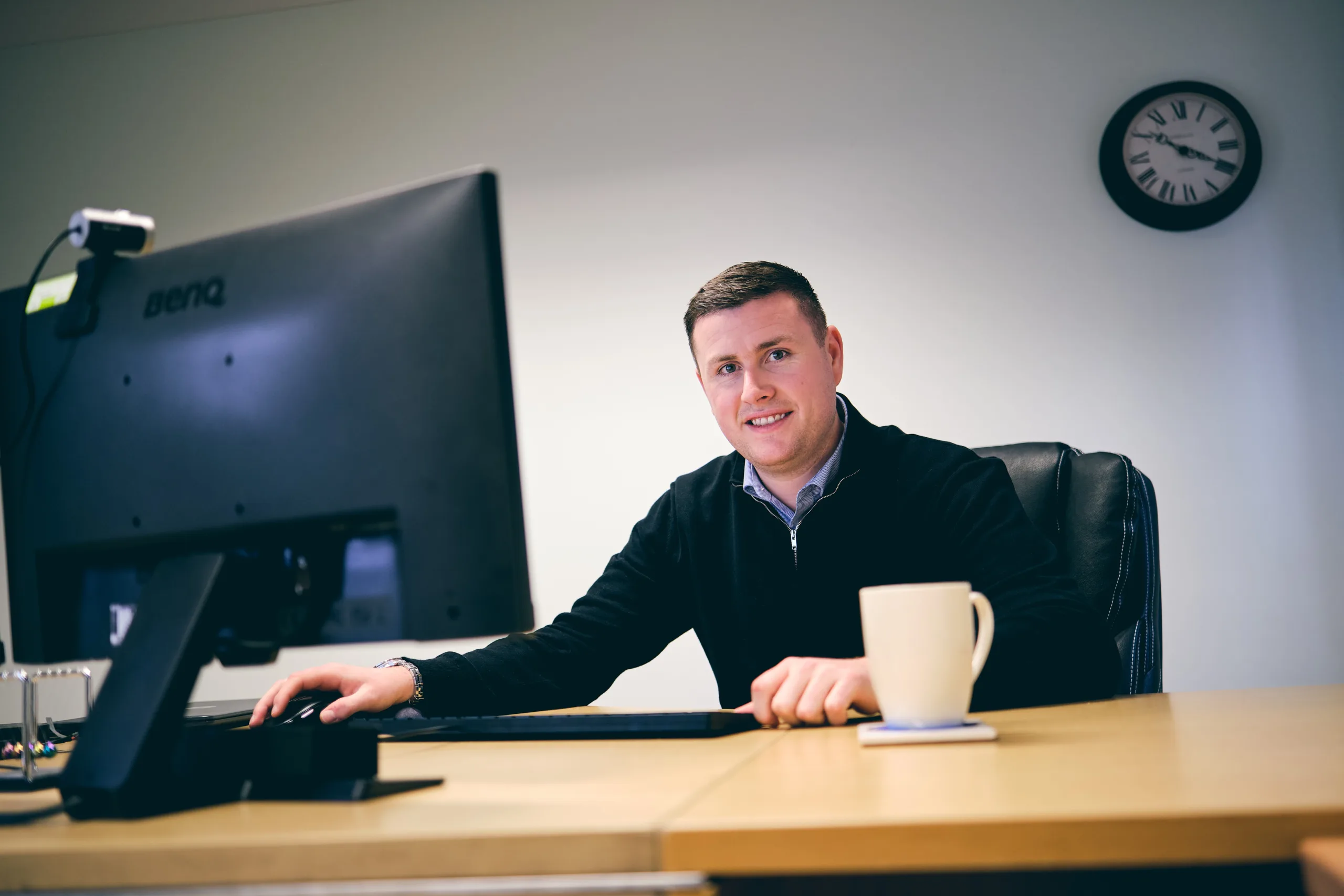 arrange your free initial phone call with one of our qualified Financial Planners, like Sam Harrington in this image. Sam sits behind his desk, smiling. He is wearing a black top and has a white mug in front of him on the desk.