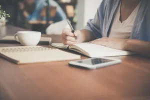 decorative image for student finance blog post. scene with person who's face isn't shown writing in a journal with their phone and laptop on the desk nearby