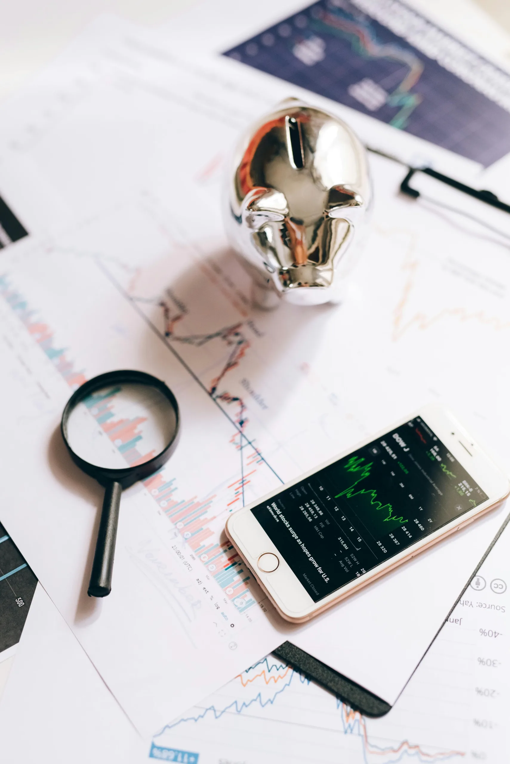 decorative image. piggy bank standing on top of papers with graphs and charts. a smartphone with charts on the screen and magnifying glass are lying nearby.