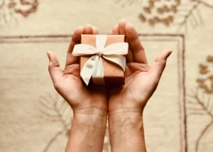 decorative image: hands holding a small gift box with a box on it. the box is pink. there is a cream coloured background under the hands. Image by Kim Stiver