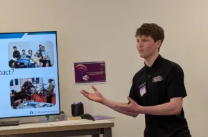 Joe from Two Wheel Campus stands in front of a TV screen showing a presentation. Social Ventures Pitch Event