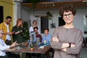 middle aged femail with short curly hair and wearing glasses stands with arms folded to the right of the image. In the background a team of people are gathered around a desk having a discussion. To accompany blog on business.