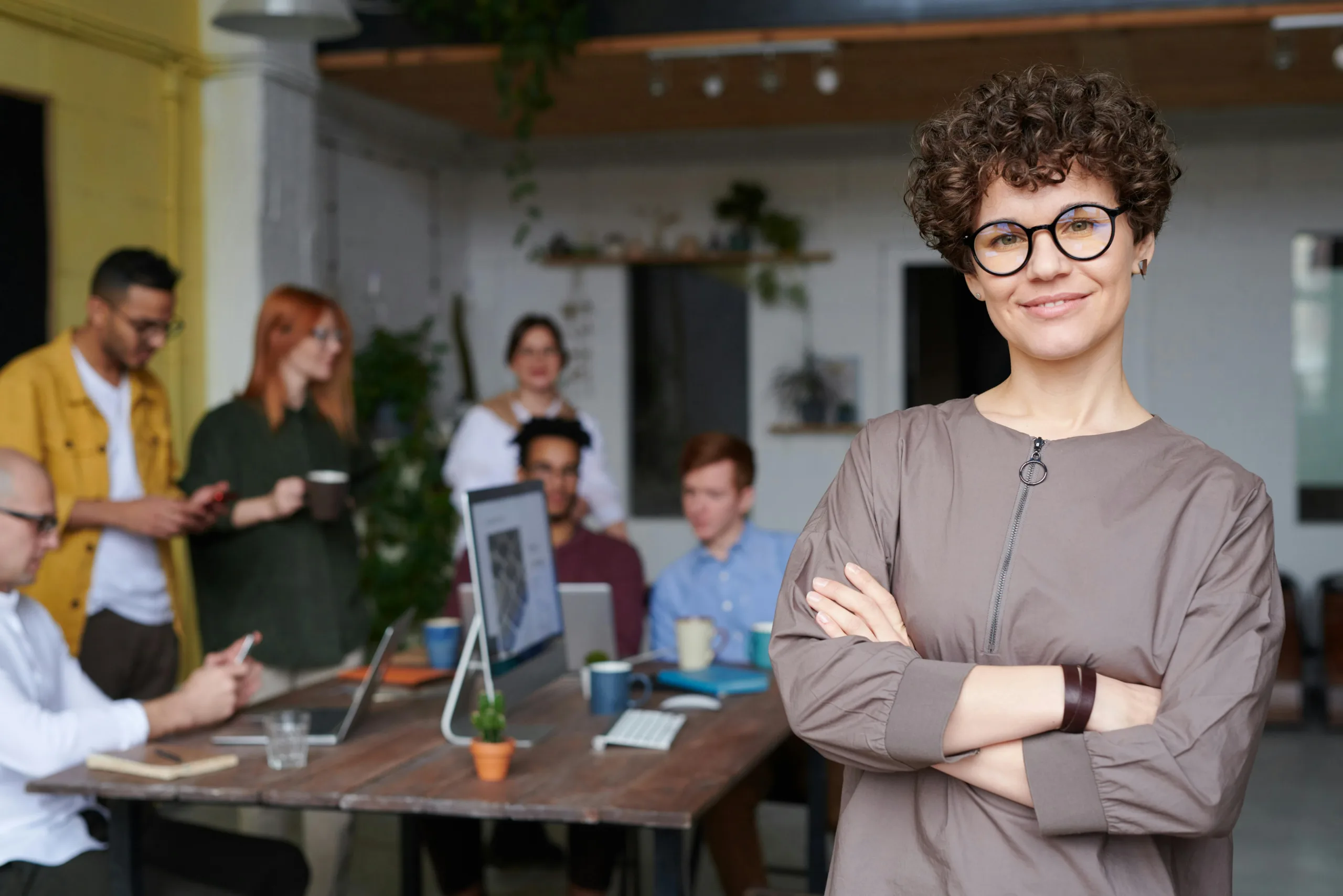 middle aged femail with short curly hair and wearing glasses stands with arms folded to the right of the image. In the background a team of people are gathered around a desk having a discussion. To accompany blog on business.