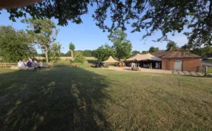 The Hall Barns at Prestwold was teh vanue for our summer BBQ. A photo of the setting showing grassed area at the front and the tent with people sitting underneath at the back.