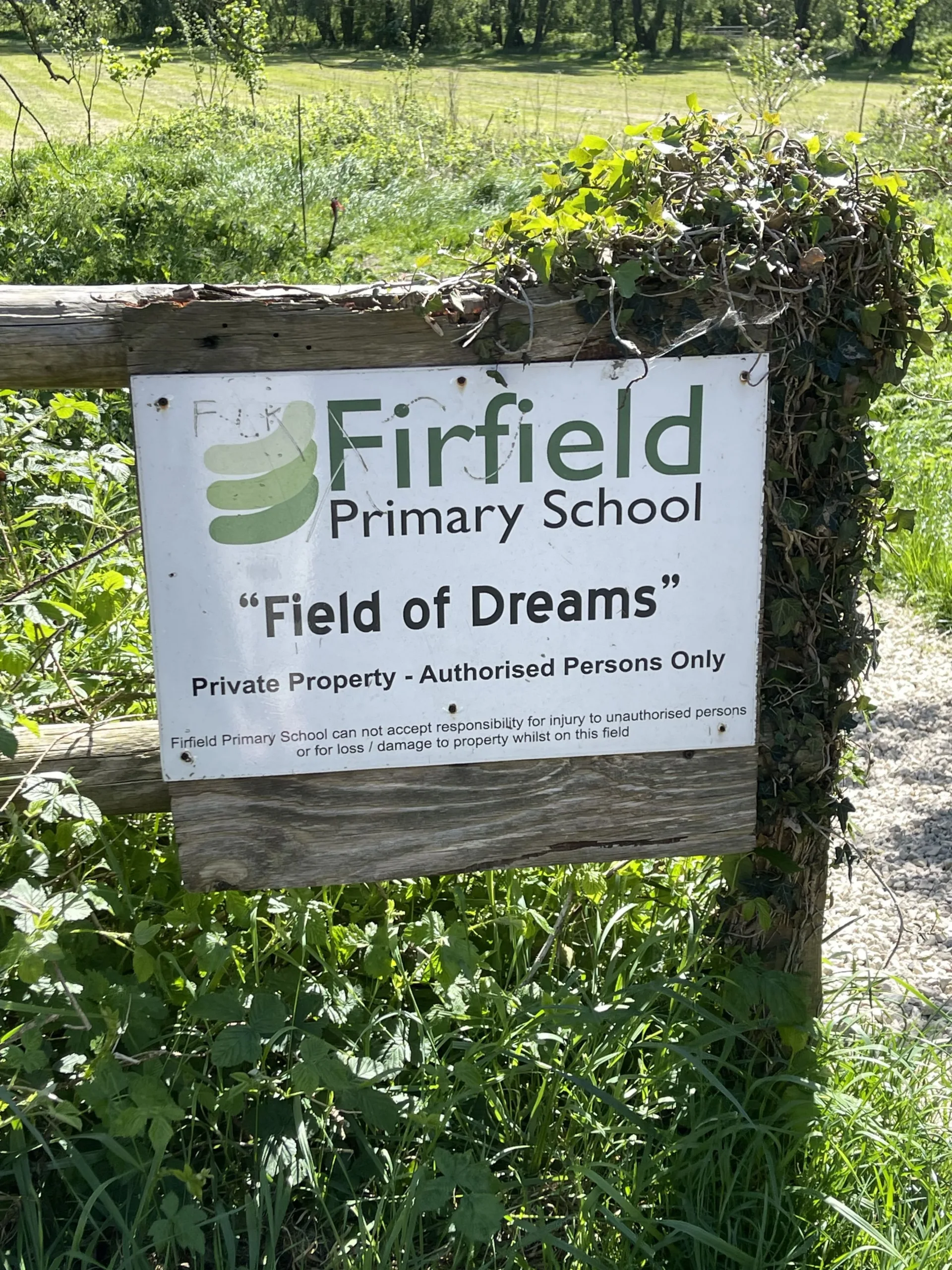 Image of a sign for Firfield Primary School on a fence with grass and bushes behind it to accompany blog post on the grant from the Chesterton House Foundation towards their allotment project